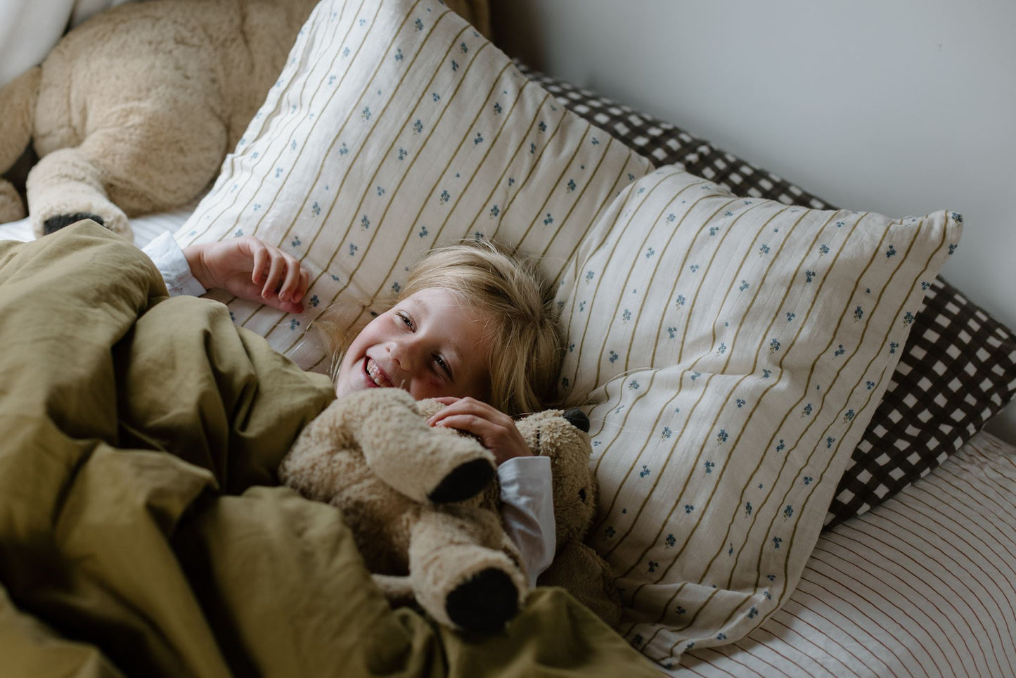 Child lying in bed with pillows and a teddy bear, surrounded by soft bedding.