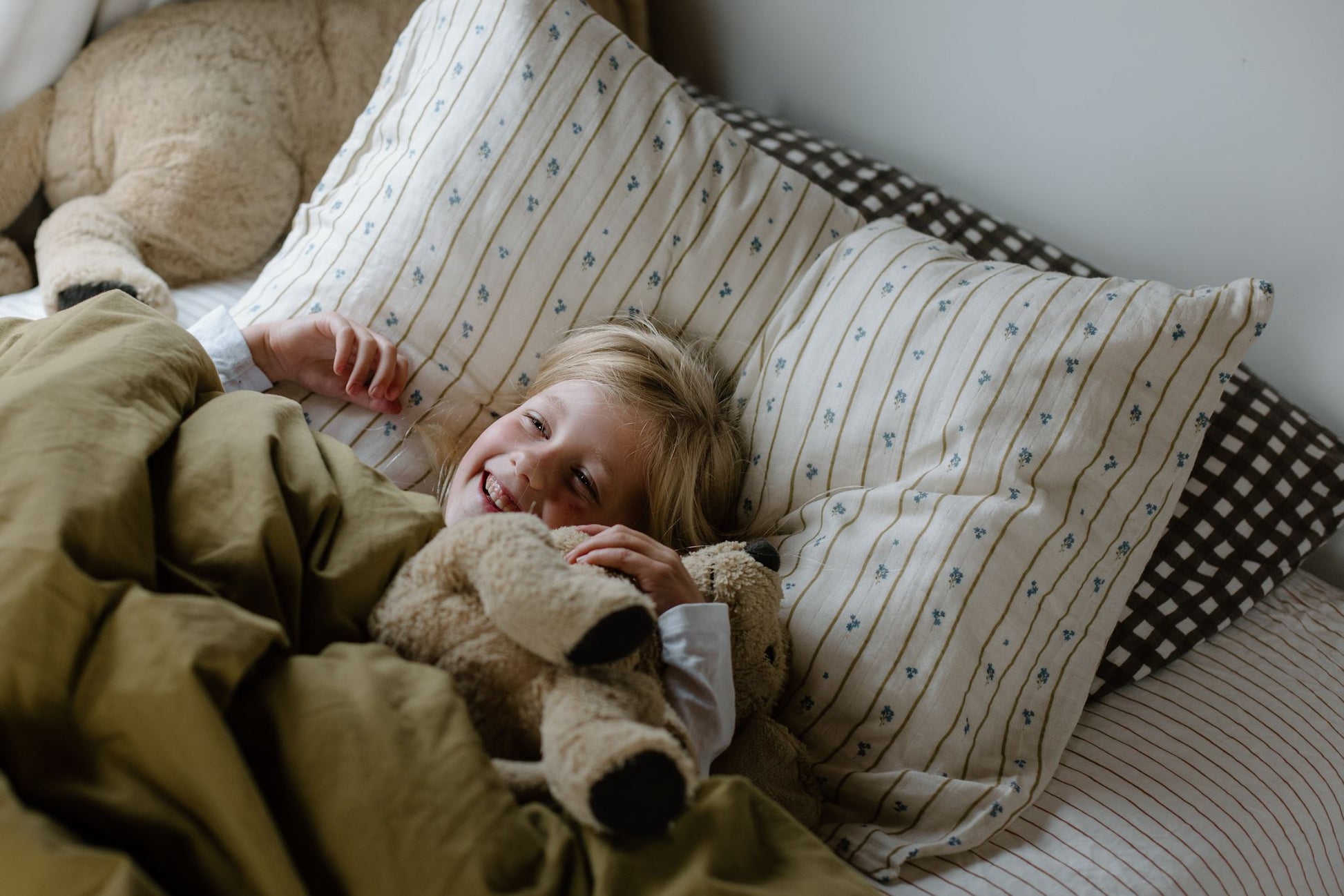 Child lying in bed with pillows and a teddy bear, surrounded by soft bedding.