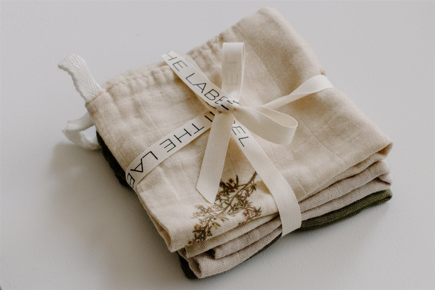 A stack of beige organic cotton muslin cloths with a brand label tied with a ribbon, designed in Wanaka, New Zealand.